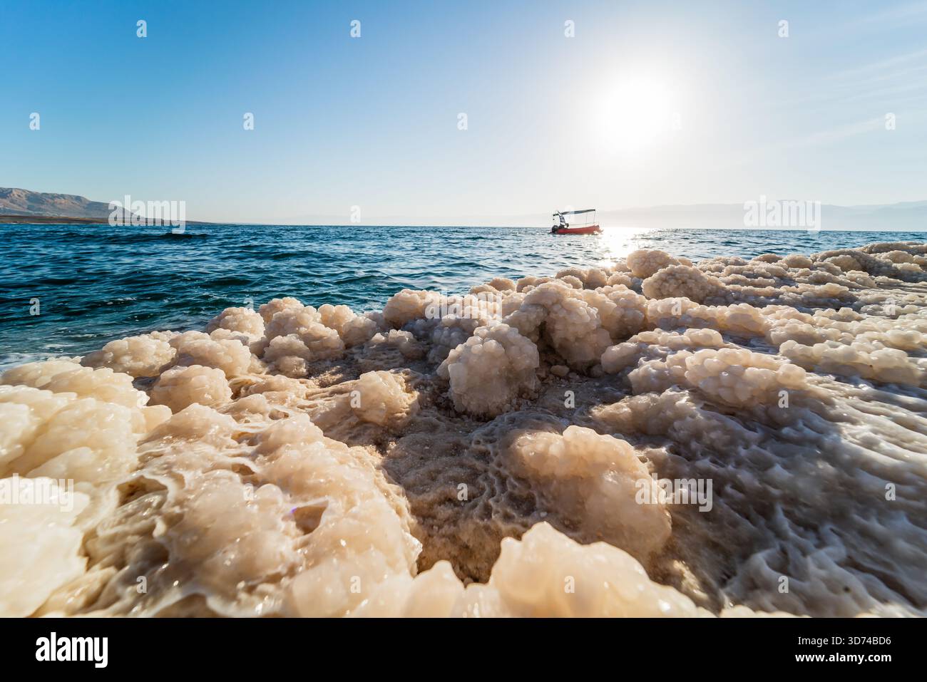 Lever de soleil sur la mer morte avec une eau turquoise calme, des dépôts de sel blanc le long du rivage, un petit bateau et un horizon lointain dans une lumière chaude. Banque D'Images