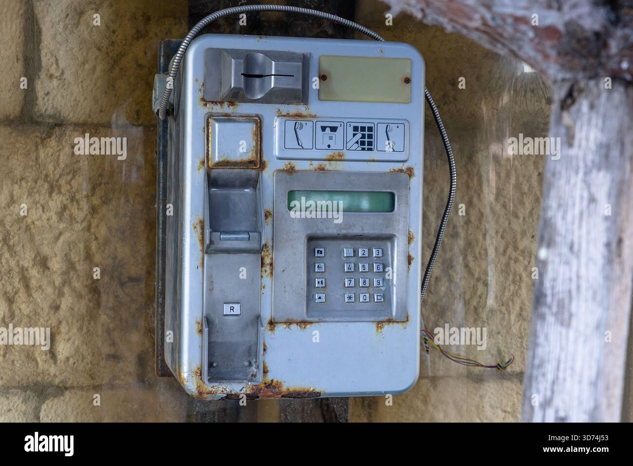 Téléphone public vieilli, moucheté par la rouille, fixé à un mur extérieur en pierre altérée. Le clavier usé et les fils lâches évoquent la nostalgie et la désintégration d'un ubiq unique Banque D'Images