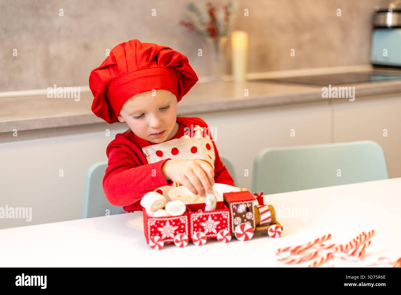 Un enfant joyeux dans un chapeau de chef rouge et un tablier place soigneusement des bonbons sur un train jouet. La cuisine est décorée pour les vacances, créant un atmos chaleureux Banque D'Images