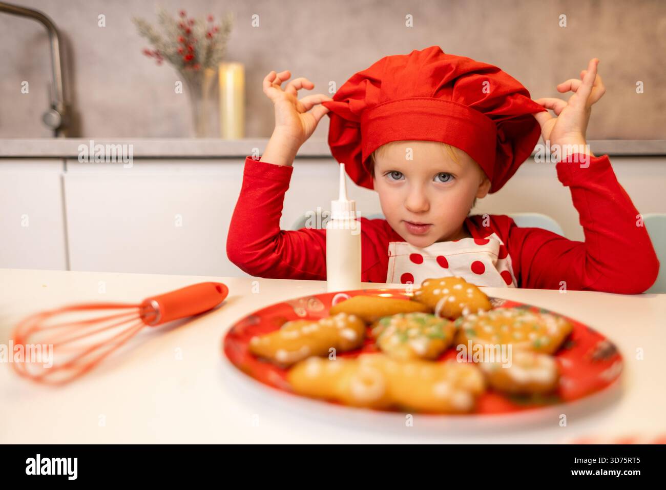 Un enfant joyeux dans un chapeau de chef rouge est assis à la table, prêt à décorer des biscuits colorés de vacances. La cuisine est lumineuse, créant une atmosphère joyeuse Banque D'Images