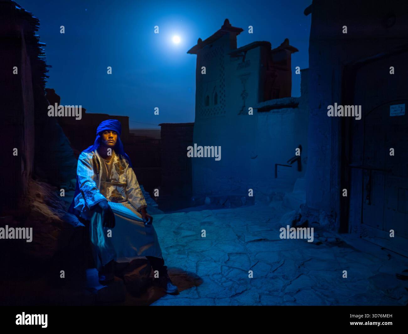 Portrait d'un homme berbère portant le turban bleu traditionnel et djellaba, assis calmement sous la pleine lune lumineuse devant les tours de kasbah en briques de boue à ni Banque D'Images