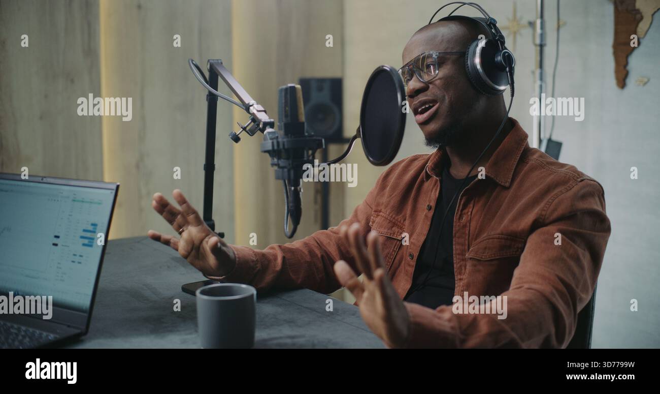 Homme adulte parle dans Professional Studio Mic avec filtre Pop, portant des écouteurs. Le client fait des gestes naturels tout en discutant des Insights, avec l'ordinateur portable affichant les analyses et la tasse à café sur la table. Banque D'Images