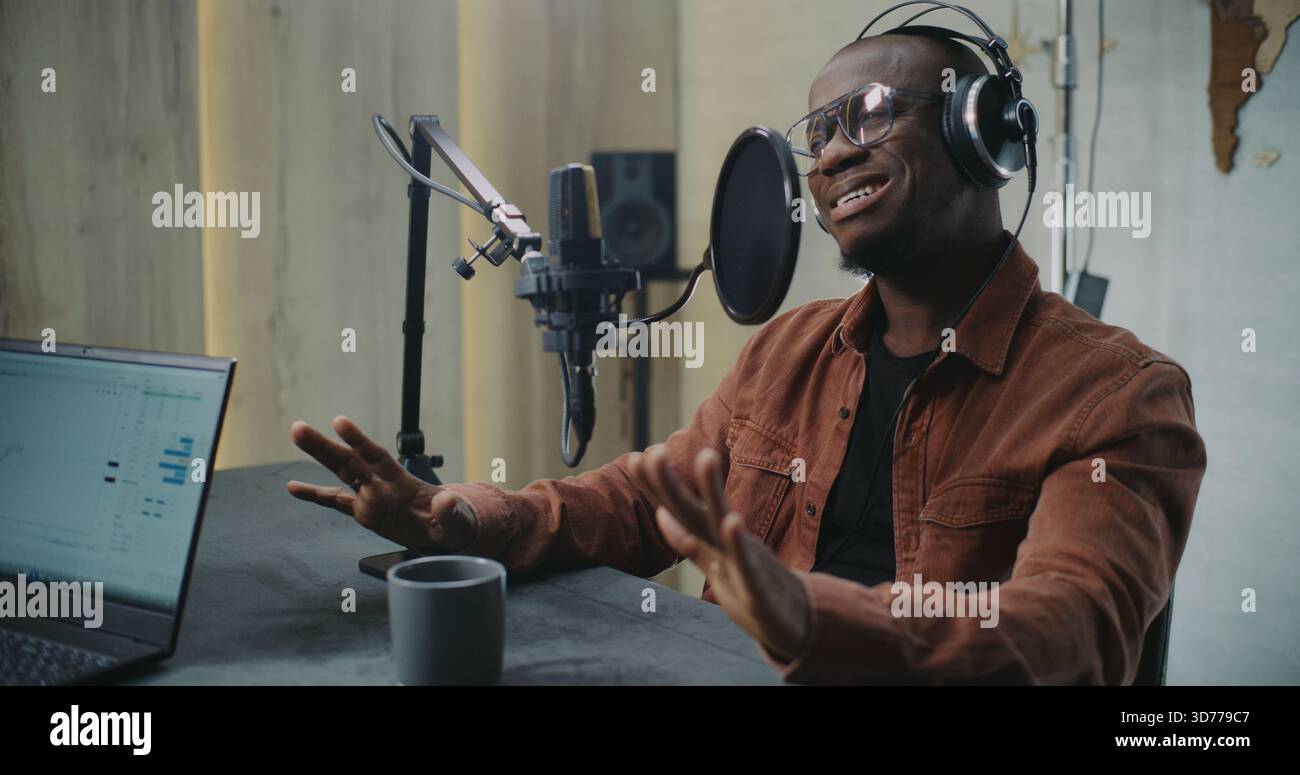 Homme adulte parle dans Professional Studio Mic avec filtre Pop, portant des écouteurs. Le client fait des gestes naturels tout en discutant des Insights, avec l'ordinateur portable affichant les analyses et la tasse à café sur la table. Banque D'Images
