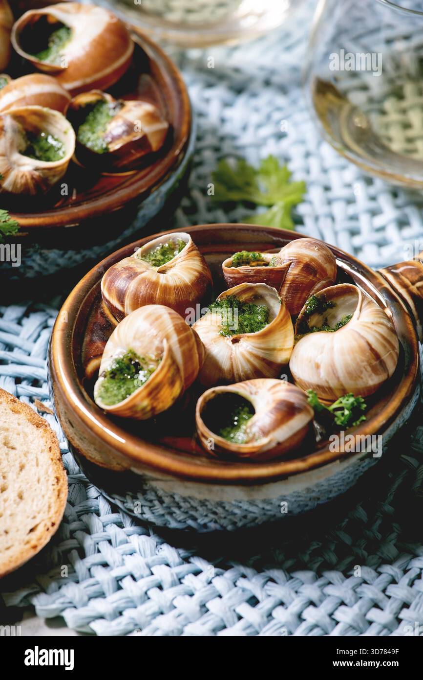 Escargots de Bourgogne - escargots aux herbes beurre, plat gastronomique, dans une casserole traditionnelle en céramique avec coriandre, pain, verre de vin blanc sur serviette de paille bleue. Gros plan Banque D'Images