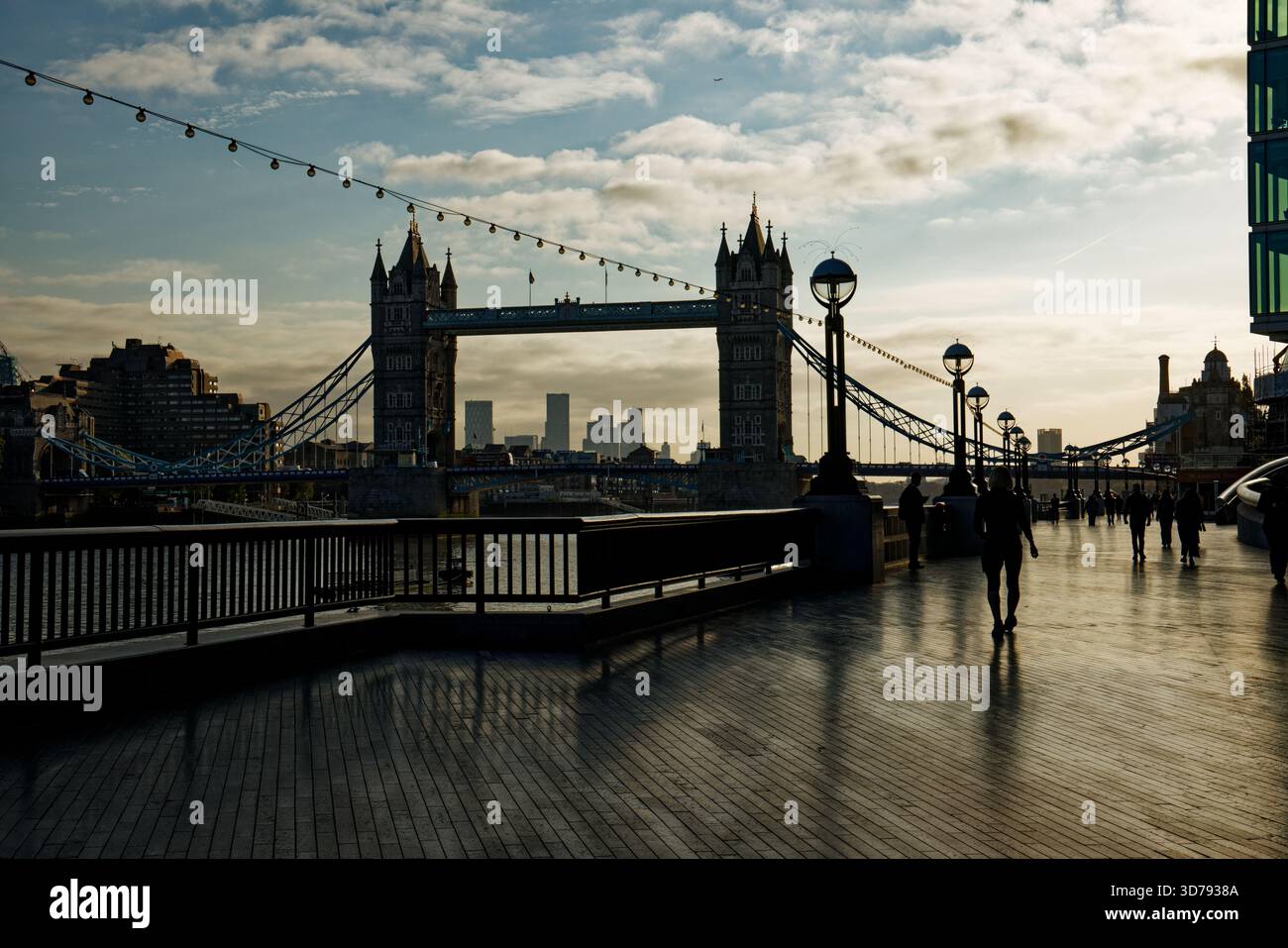 Les gens sont silhouettés dans l'éblouissement de la lumière du soleil du matin comme la promenade le long de la rivière de la Tamise avec Tower Bridge en arrière-plan, Londres. Banque D'Images
