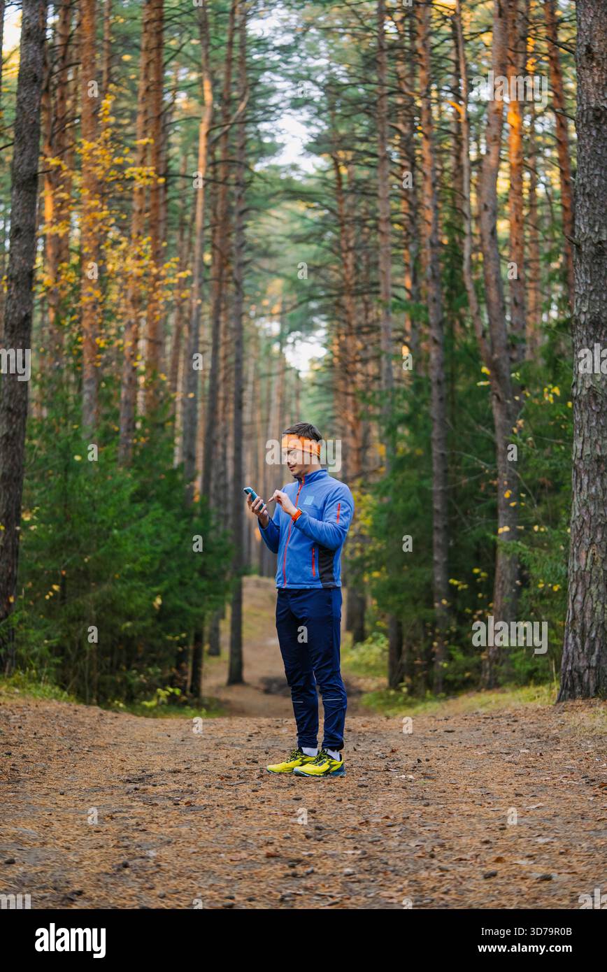 Homme portant des vêtements de sport et un bandeau orange debout sur un chemin de forêt de pins, tenant et regardant vers le bas son smartphone, éventuellement en vérifiant une carte ou une application de fitness Banque D'Images