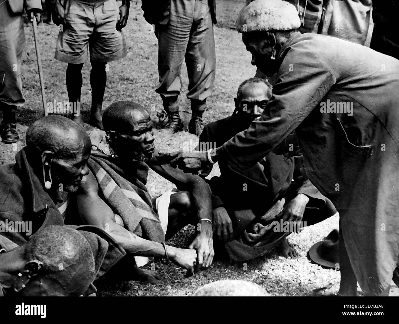 Cérémonie de nettoyage Mau-Mau -- il s'agit de la première photo prise d'une cérémonie de nettoyage Mau-Mau dans le district de Kiambu au Kenya. Sur la droite en pelage de peau de mouton, le %Mundo Mu Gu' (Un sorcier qui pratique pour le bien, pas le mal), se prépare à absoudre les membres de la tribu Kikuyu qui ont prêté le serment Mau-Mau. En face du avec docteur est une dépression dans le sol bordée de feuilles de banane, contenant le sang et le contenu de l'estomac d'une chèvre abattue à cet effet. Le sabot de la chèvre, enveloppé dans des feuilles, est plongé dans le sang et s'applique à la langue initiée, qui retape alors le ma Banque D'Images