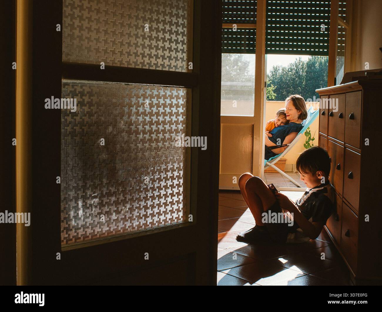 Une mère est assise dans une chaise confortable sur le balcon, tenant son enfant endormi. Un autre enfant est assis sur le sol à l'intérieur, lisant un livre. La photo capture f Banque D'Images