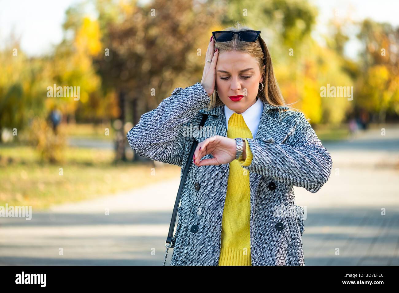 Femme d'affaires stressée et inquiète en manteau d'automne vérifiant l'heure sur sa montre-bracelet tout en se tenant dans le parc de la ville. Banque D'Images