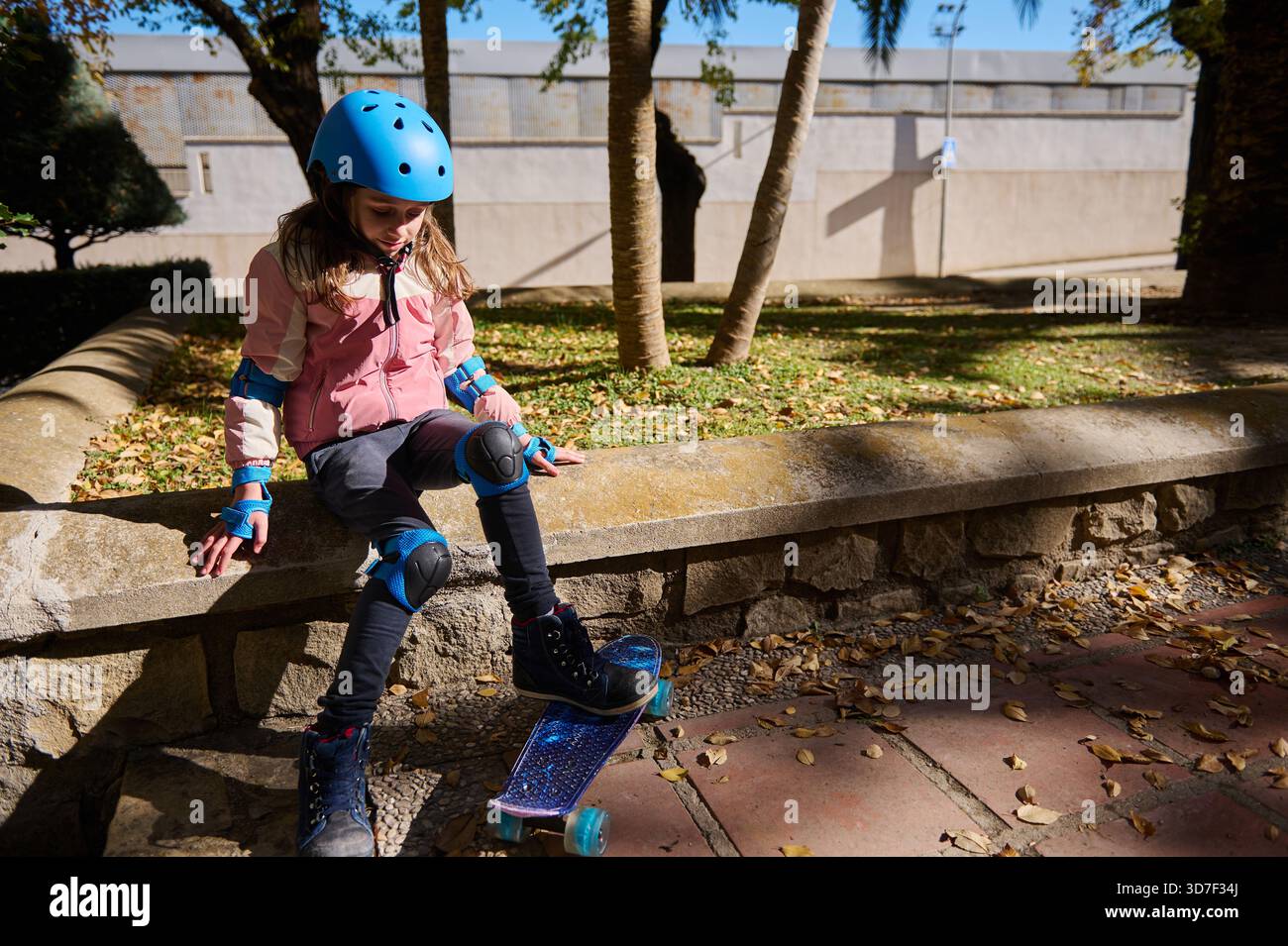 Une jeune fille porte un casque bleu et des coussinets de protection tout en s'équilibrant sur une planche à roulettes à côté d'un mur de pierre dans un parc ensoleillé. Les feuilles d'automne brillantes se dispersent Banque D'Images