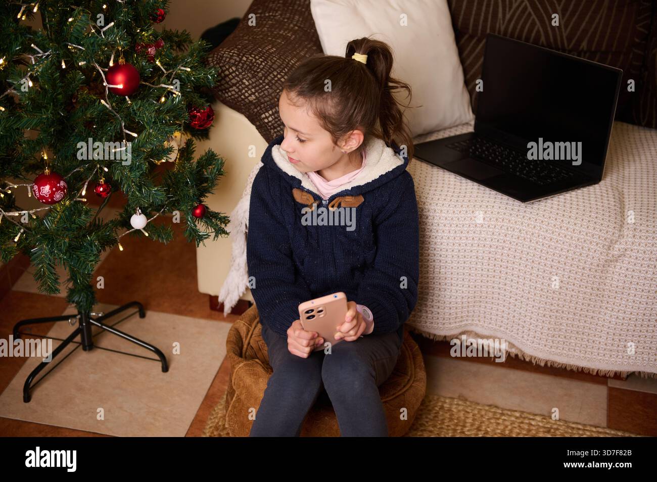 Une jeune fille est assise à côté d'un arbre de Noël décoré dans un salon confortable, tenant un téléphone tandis qu'un ordinateur portable repose sur le canapé, capturant des vacances chaudes Banque D'Images