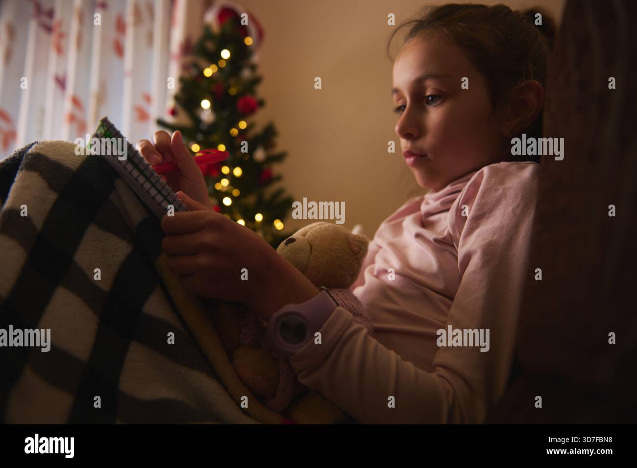 Une jeune fille est assise sous une couverture chaude, lisant un cahier tout en tenant un ours en peluche. Des lumières de Noël douces et un arbre créent un fami calme et confortable Banque D'Images