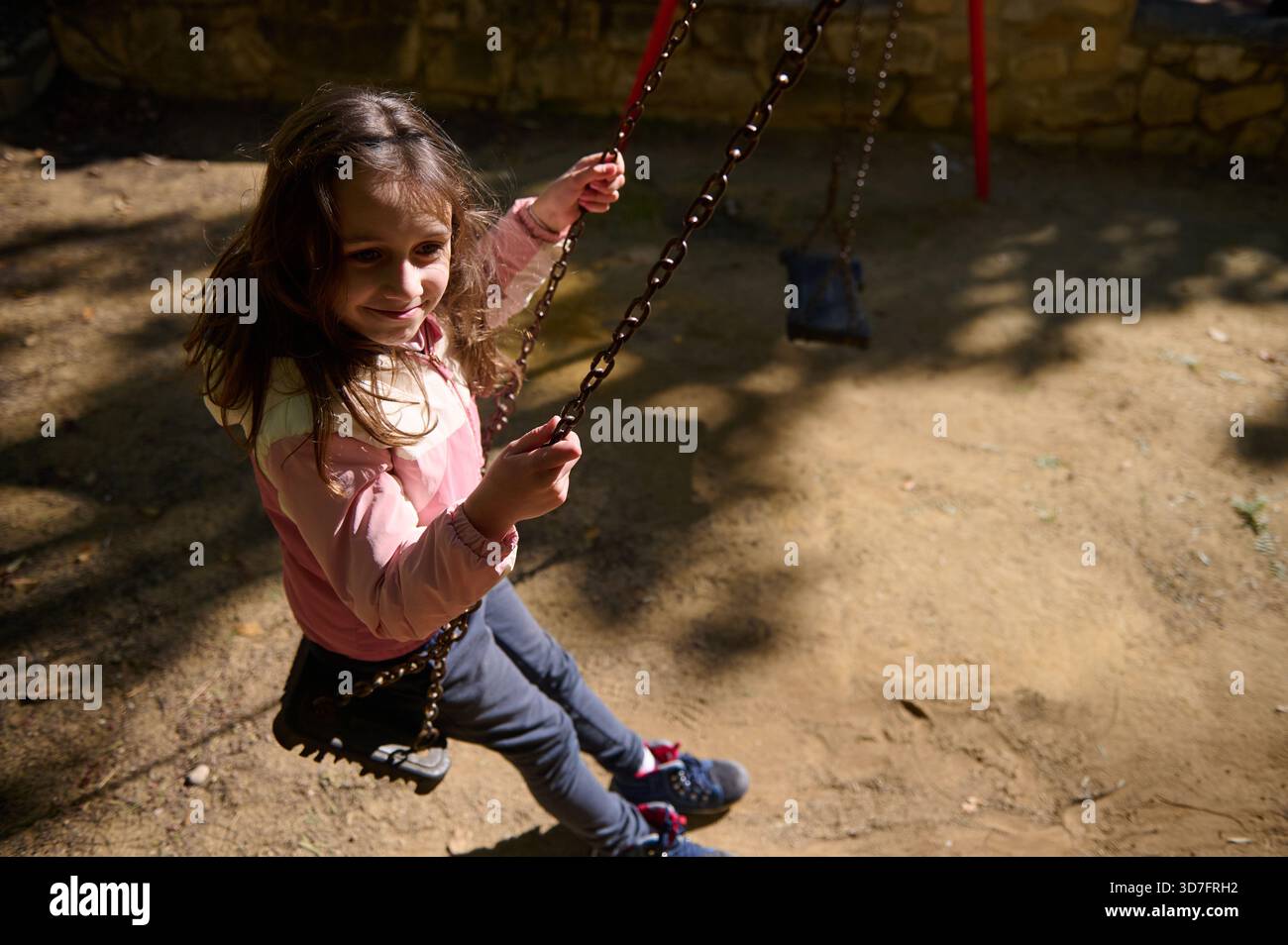 Une jeune fille dans une veste rose sourit en se balançant dans un parc ensoleillé. La scène transmet la joie de l'enfance insouciante et le jeu en plein air. Banque D'Images