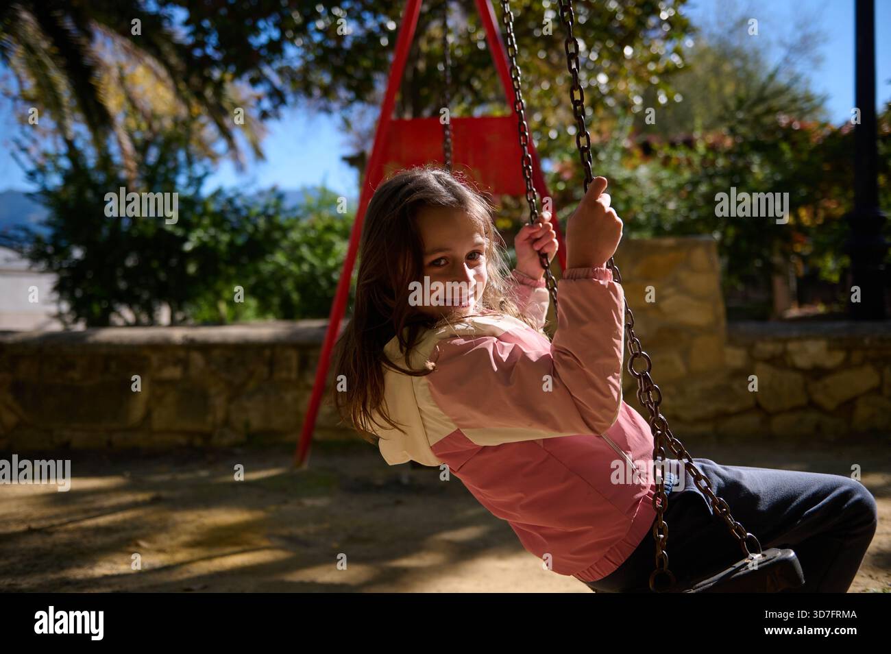 Une jeune fille souriante aime une balançoire dans un parc ensoleillé, capturant des moments d'enfance ludiques et des divertissements en plein air. Banque D'Images