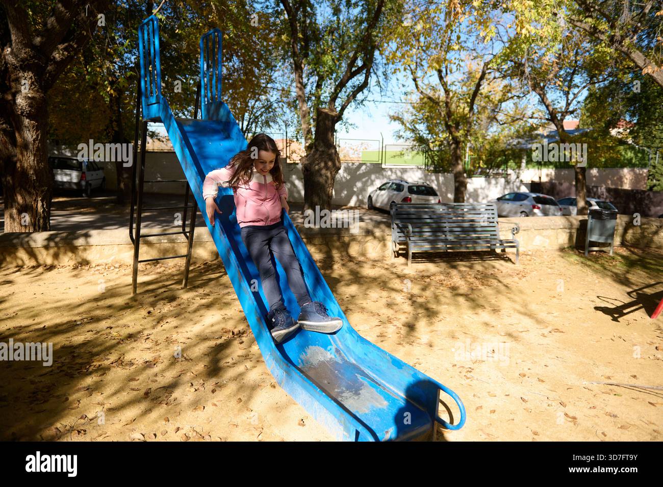 Une jeune fille dans un sweat à capuche rose glisse le long d'un toboggan bleu vif de terrain de jeu tandis que la lumière du soleil filtre à travers les arbres. Un banc de stationnement et des voitures garées créent un chee Banque D'Images