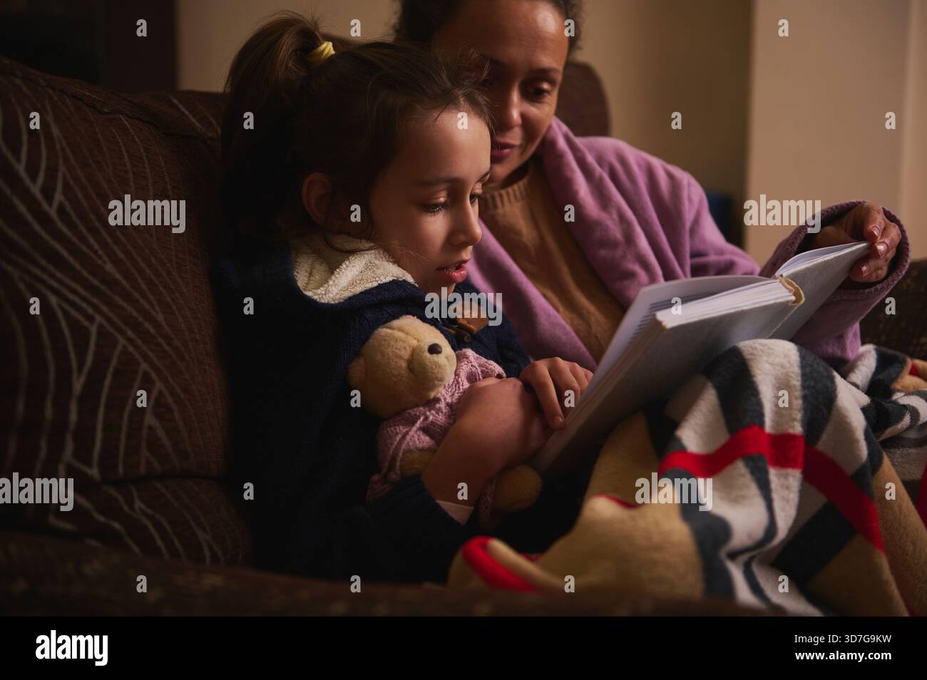Une scène tendre d'une mère lisant à sa fille sur un canapé, enveloppée dans une couverture rayée avec un ours en peluche à proximité, partageant un moment chaud et calme Banque D'Images