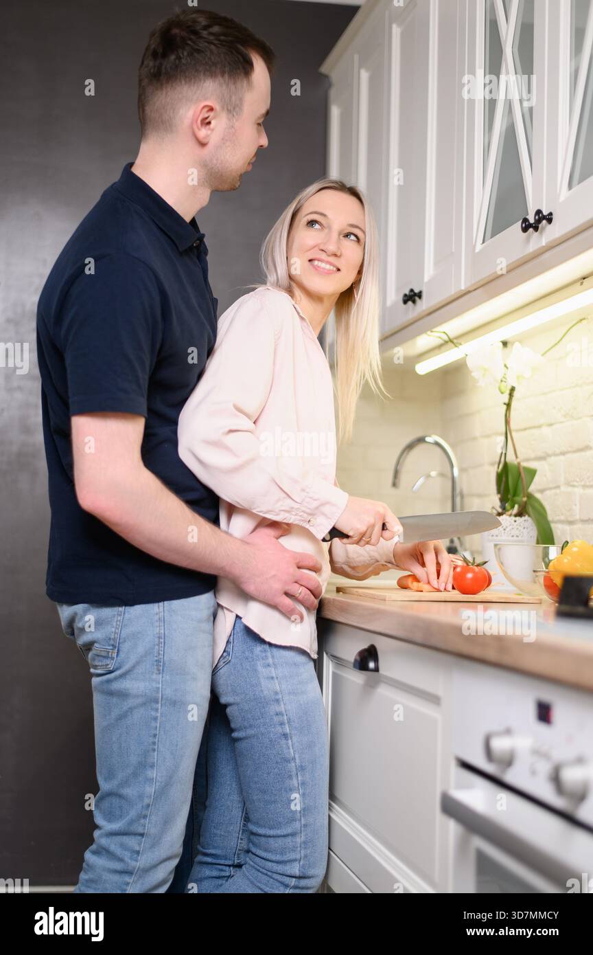 Couple cuisiner ensemble, profiter du temps dans la cuisine à la maison pendant l'escapade de week-end. Femme, amoureuse, hache des légumes dans la cuisine, tandis que l'homme l'embrasse de Be Banque D'Images