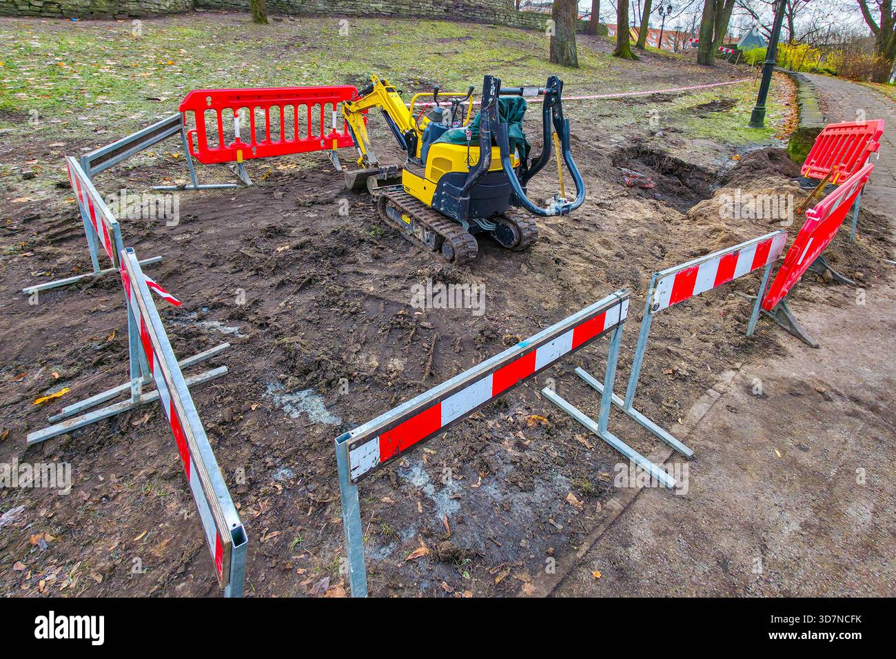 Une petite mini-pelle jaune travaille dans une zone de construction clôturée dans un parc, entourée de barrières de sécurité rouges pendant l'excavation du sol Banque D'Images