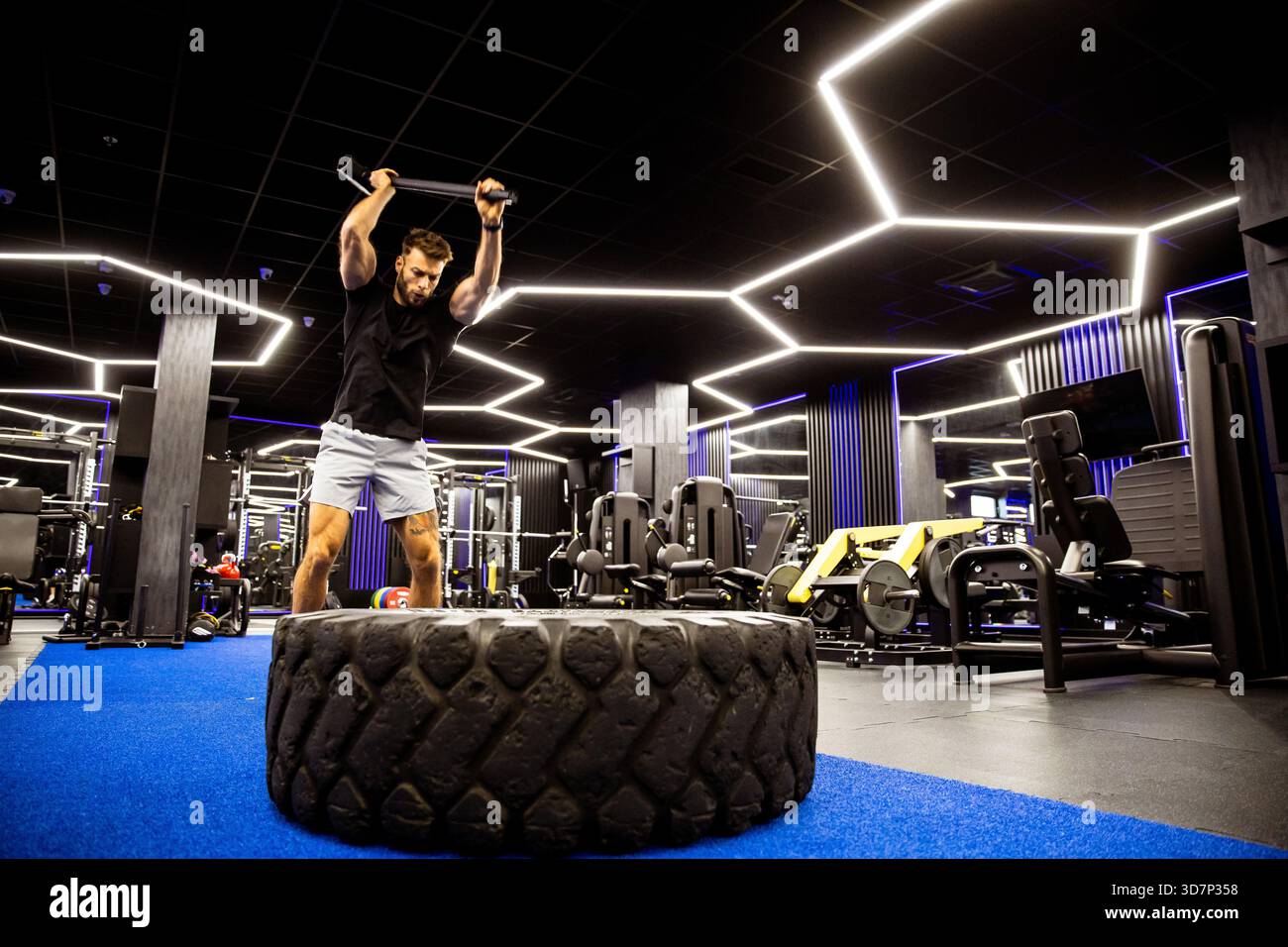 Un homme s'entraîne intensément dans un gymnase élégant, frappant un gros pneu avec un marteau. Les lumières vives et l'équipement créent une atmosphère énergisante pour h. Banque D'Images