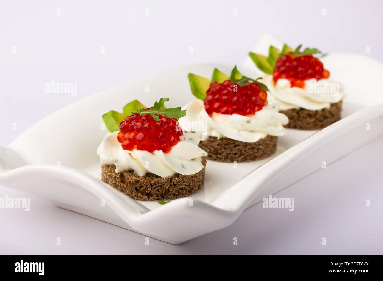 Trois délicieux canapés sont disposés sur un plat blanc avec une croûte de pain noir surmontée de crème, de caviar et d'herbes fraîches. Parfait pour n'importe quel festival Banque D'Images