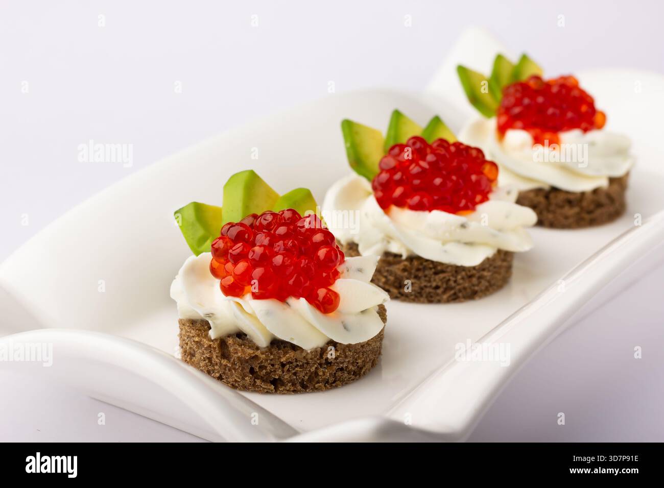 Trois canapés recouverts de fromage à la crème et de caviar rouge vif sont élégamment posés sur un plat de service blanc, garni de tranches d'avocat. Banque D'Images