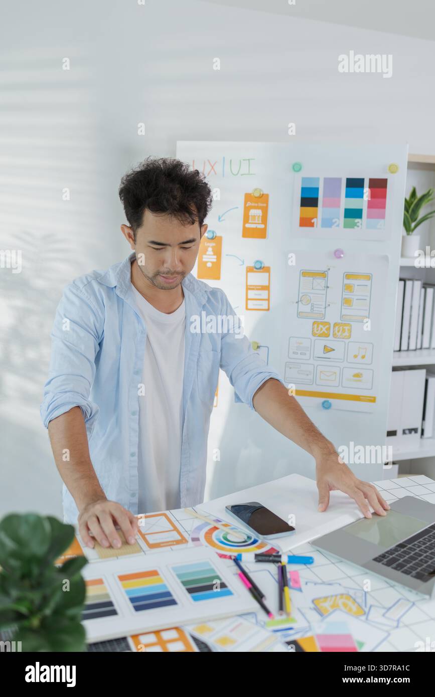 Un concepteur créatif travaille sur la planification UX/UI, analysant des croquis et des prototypes sur un tableau blanc tout en utilisant un ordinateur portable. Le processus montre la pensée de conception Banque D'Images