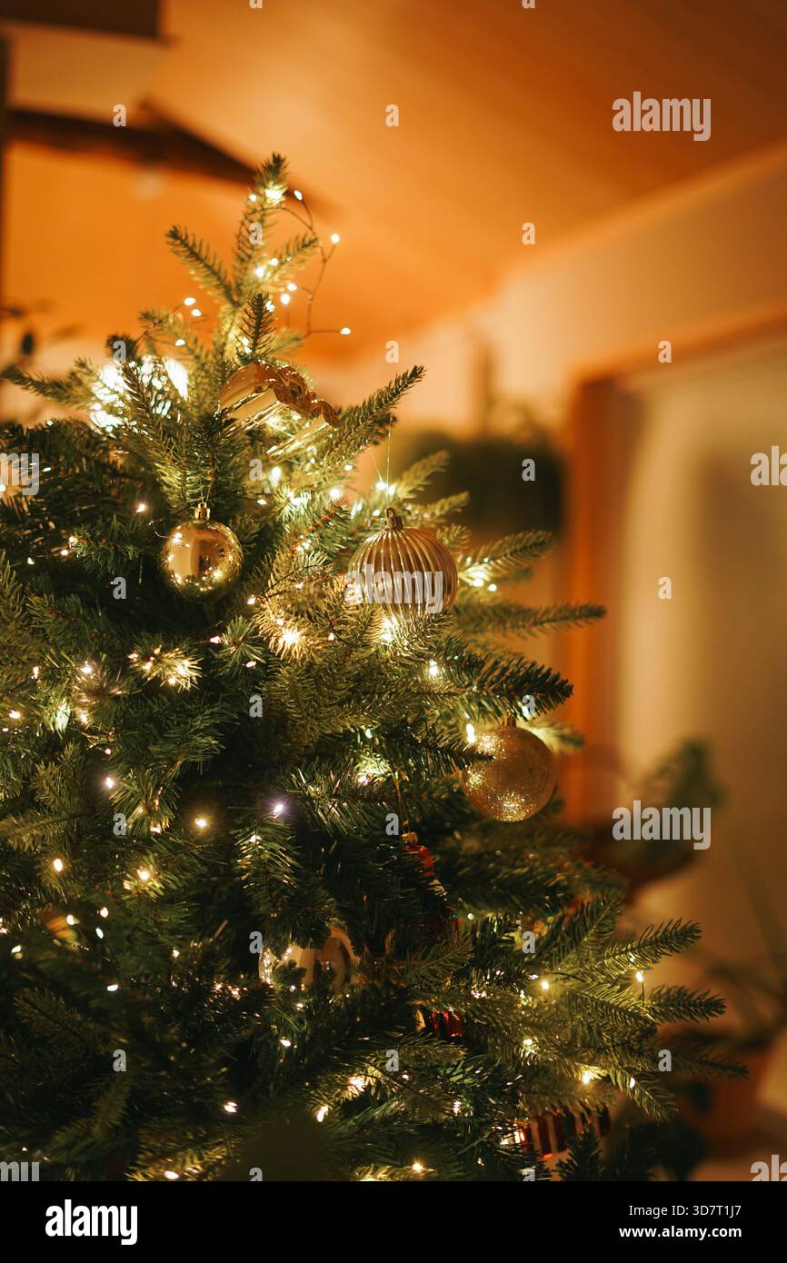 Gros plan des ornements dorés et des lumières scintillantes sur un sapin de Noël intérieur avec éclairage orange chaud Banque D'Images