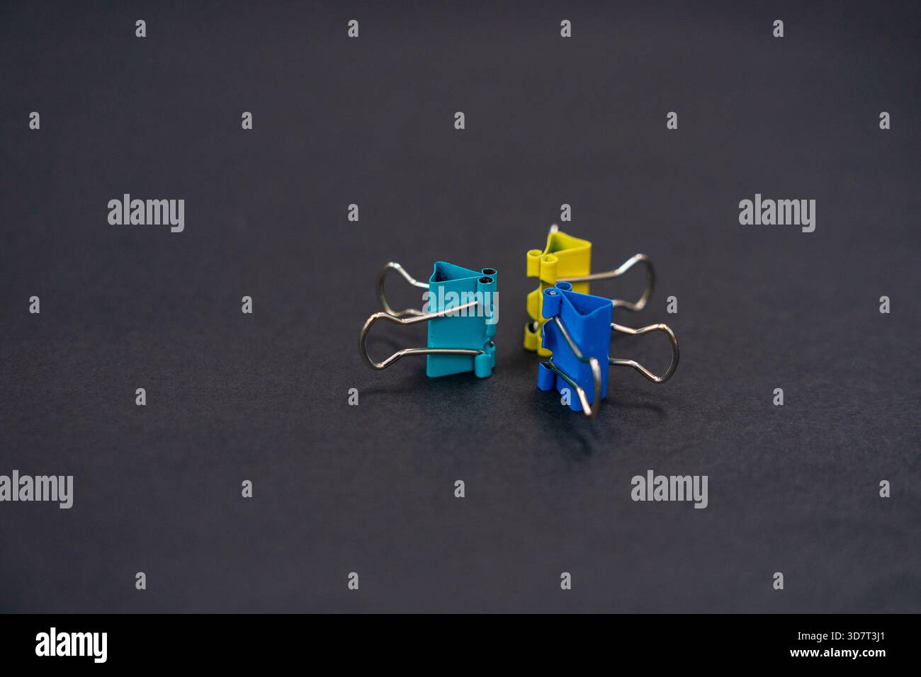 Une vue rapprochée de trois pinces de reliure colorées (sarcelle, jaune et bleue) debout sur une surface texturée gris foncé. Banque D'Images