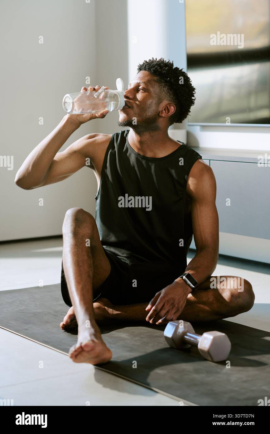 Jeune homme noir adulte assis sur le tapis de yoga eau potable après l'entraînement matinal Banque D'Images