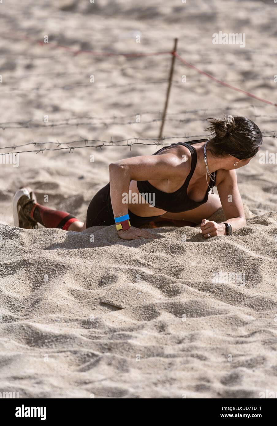 Gros plan frontal depuis le niveau du sol d'une athlète déterminée rampant dans le sable profond sous un obstacle de fil de fer barbelé, regardant en arrière pour voir s'il y en a un Banque D'Images
