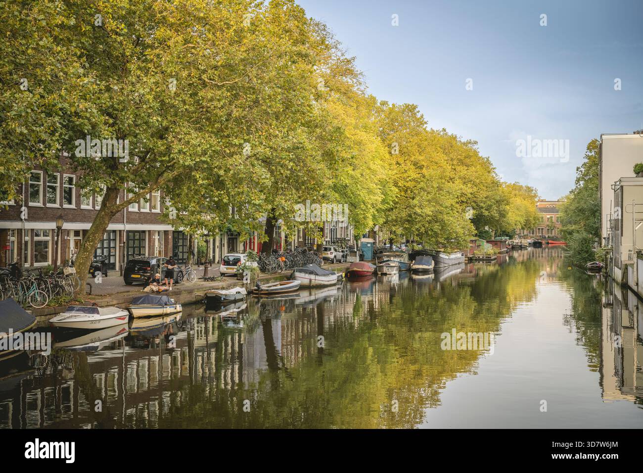 Canal bordé d'arbres avec des bateaux, des reflets et des bâtiments historiques sous un ciel bleu clair. Amsterdam,. Pays-Bas Banque D'Images
