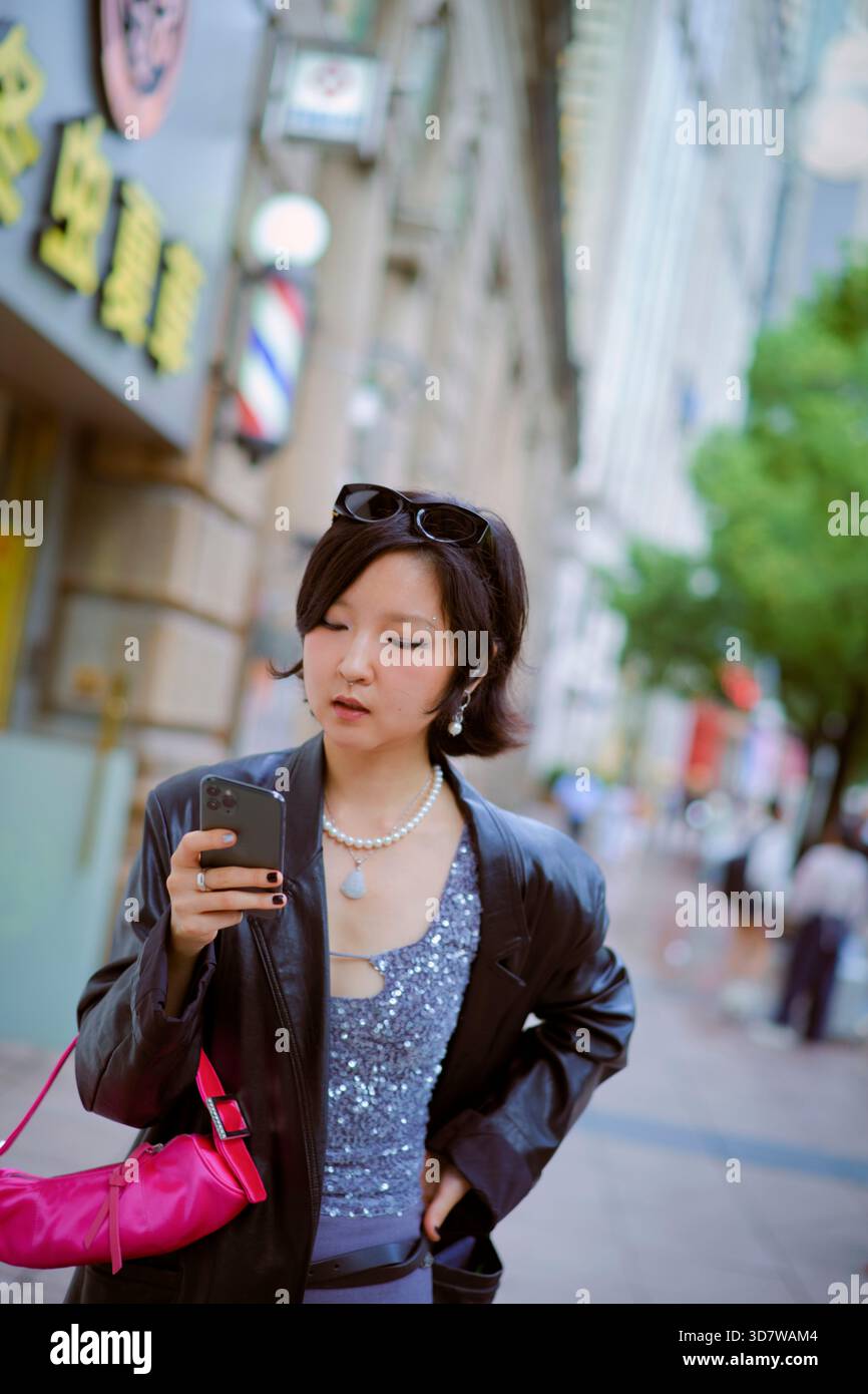 Jeune femme dans une veste en cuir vérifiant le téléphone sur la rue animée de la ville. Shanghai, Chine Banque D'Images