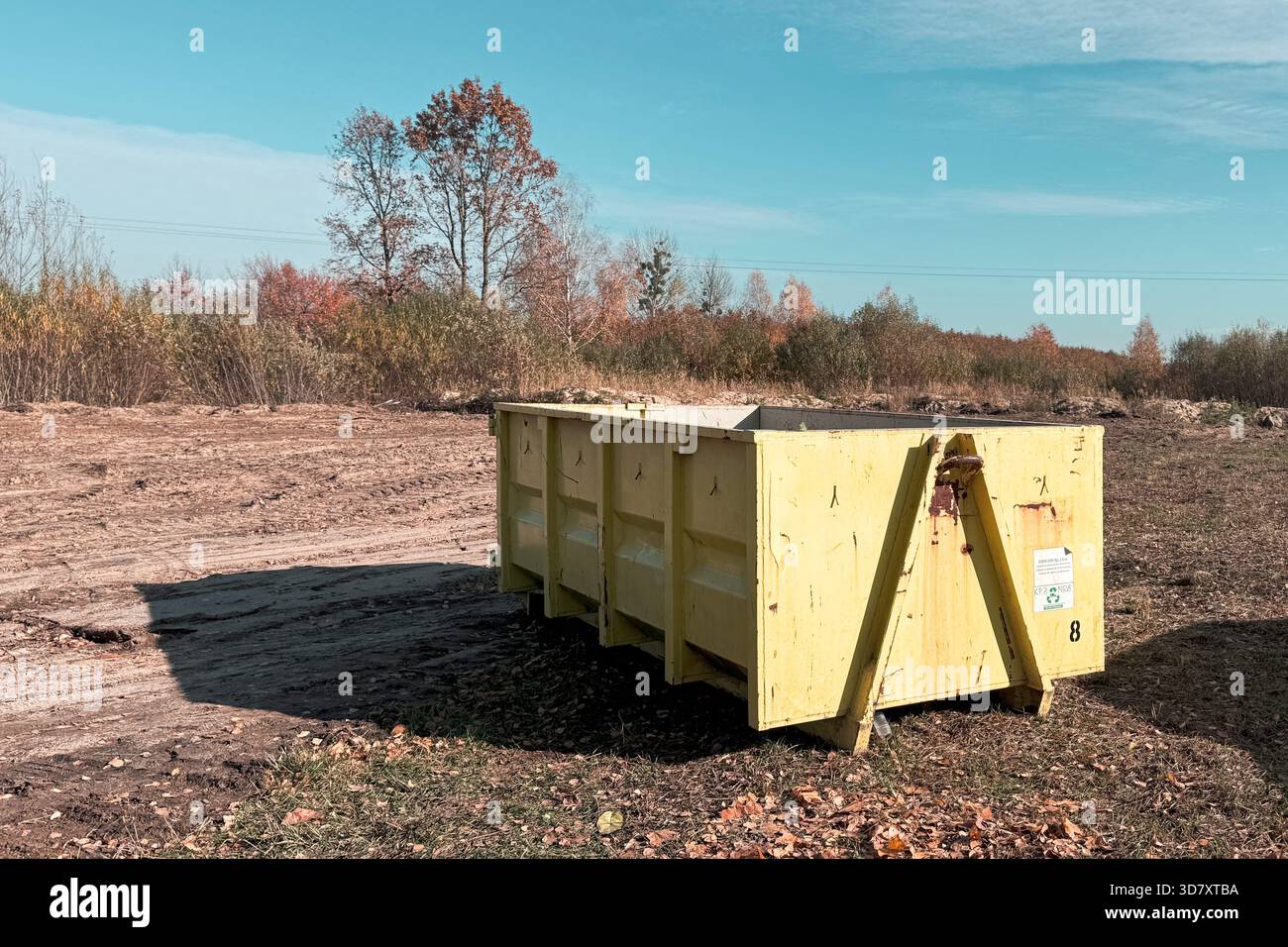 Grand conteneur de déchets en métal jaune debout sur un champ de terre dégagé avec des arbres aux couleurs d'automne en arrière-plan. Banque D'Images