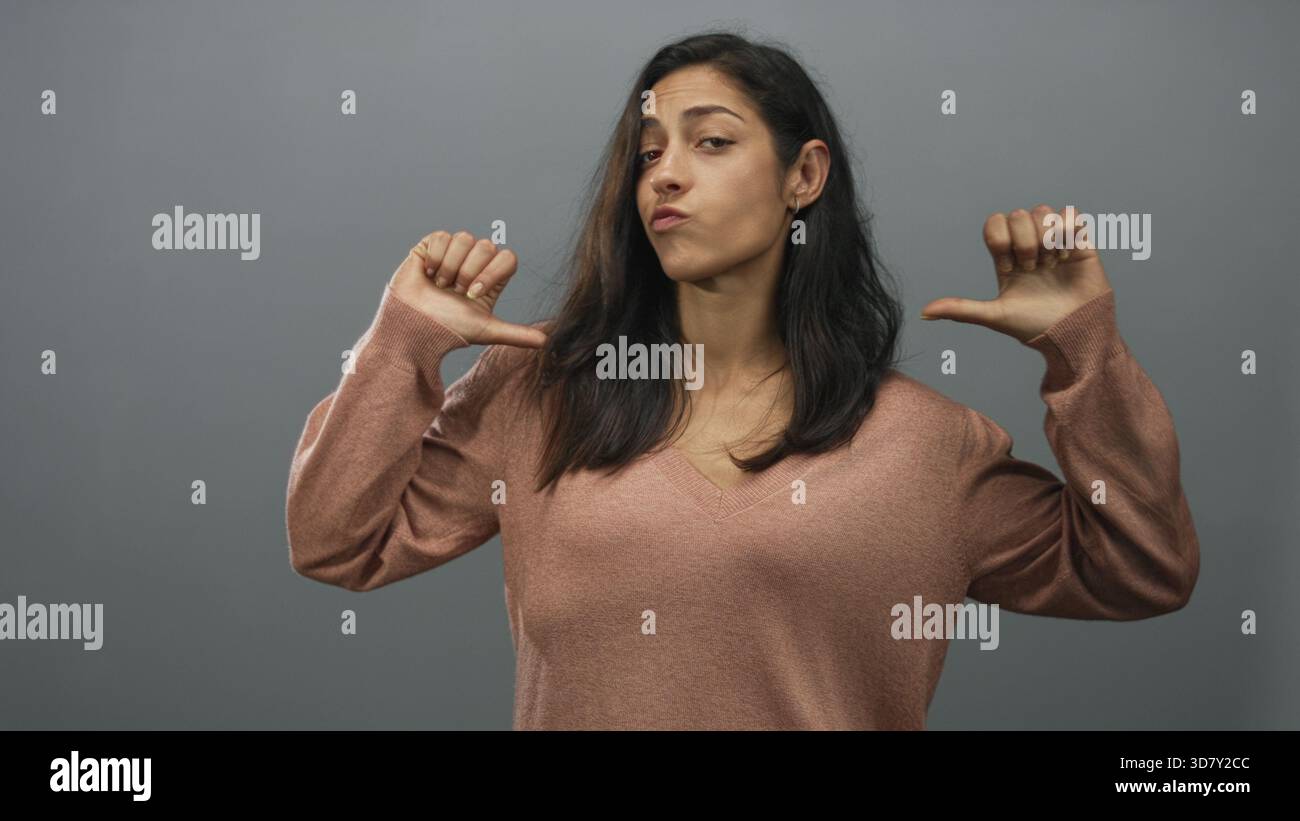 Femme avec les pouces pointant vers la poitrine dans un studio gris portant un pull à col v rose et les yeux fermés, pose décontractée ; attitude sarcastique. Banque D'Images