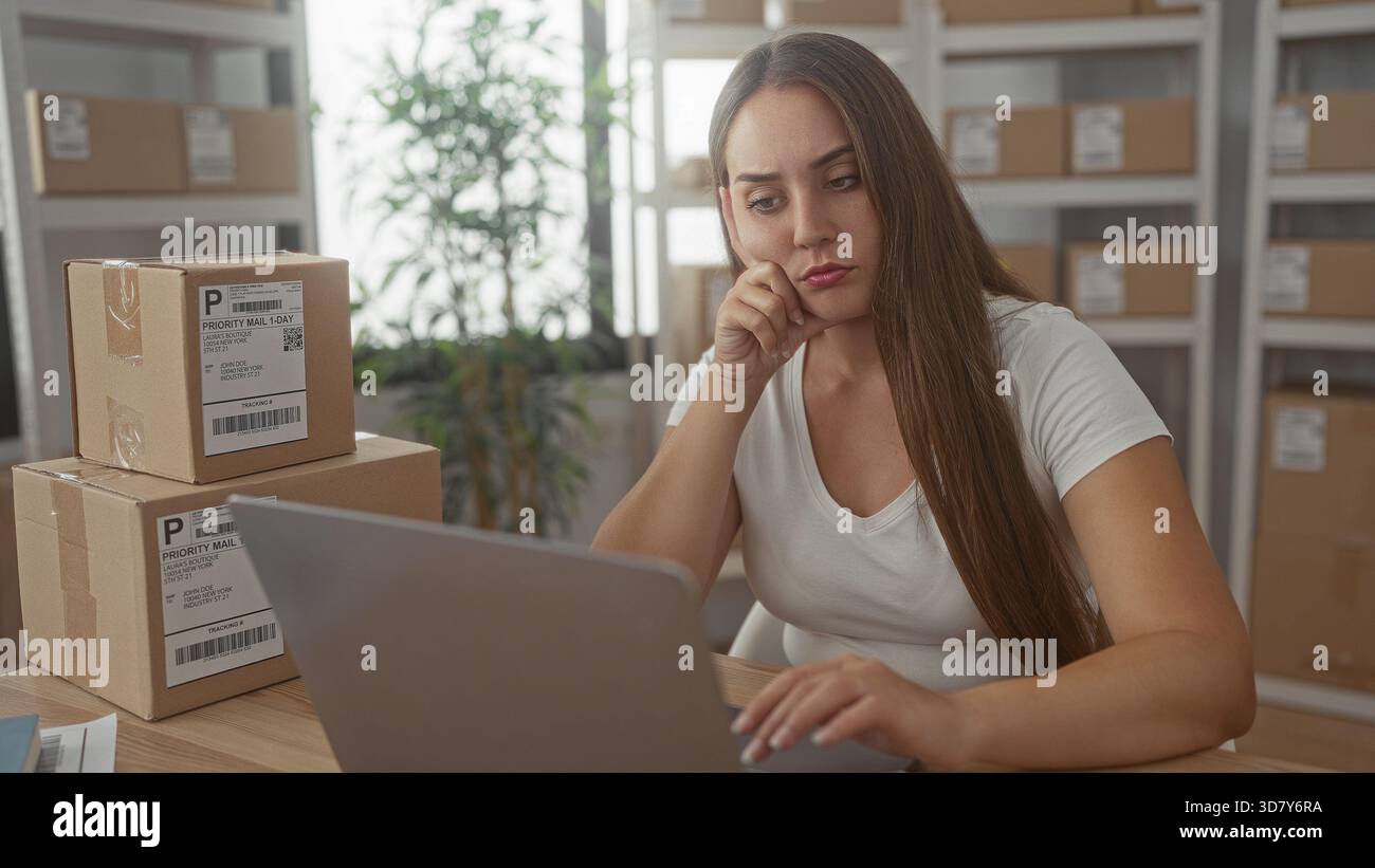 Femme tapant sur ordinateur portable à côté de colis empilés traitant les commandes en ligne dans l'immeuble de bureaux ; planification pensive de petite entreprise. Banque D'Images