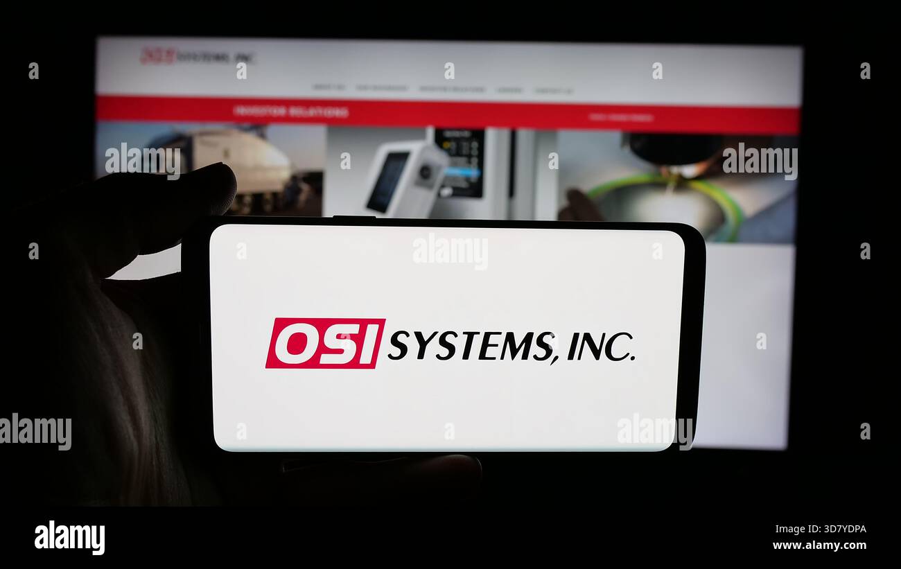 Stuttgart, Allemagne - 11-18-2025 : personne tenant un téléphone portable avec le logo de la société de sécurité américaine OSI Systems Inc sur l'écran devant la page Web. Banque D'Images