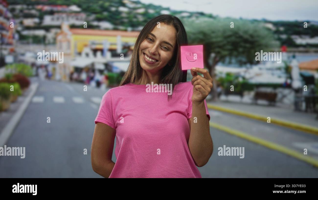 Femme tenant une note collante rose avec un visage souriant dans la rue souriant et montrant le visage tout en portant un tshirt rose ; positivité de joie. Banque D'Images