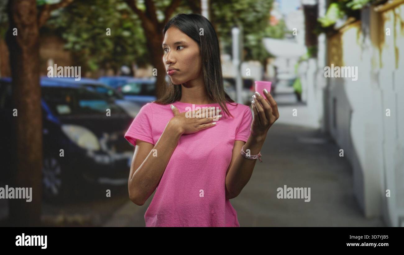 Femme thaïlandaise tient la coupe menstruelle rose dans une main et place l'autre main à la poitrine dans une rue urbaine étroite avec des voitures garées ; hésitation curiosité. Banque D'Images