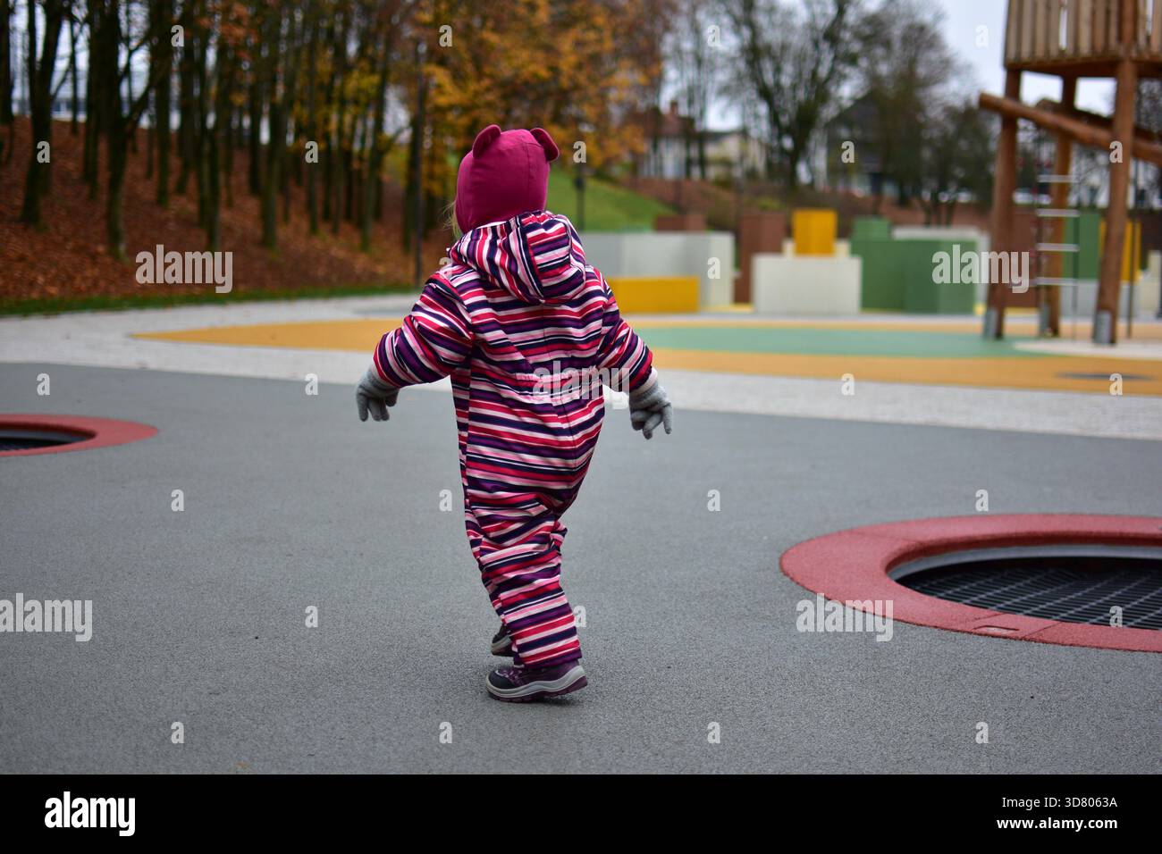 Jeune fille dans un hiver rayé marchant en toute confiance à travers une aire de jeux extérieure moderne avec un sol en caoutchouc brillant et des structures de jeu en bois Banque D'Images