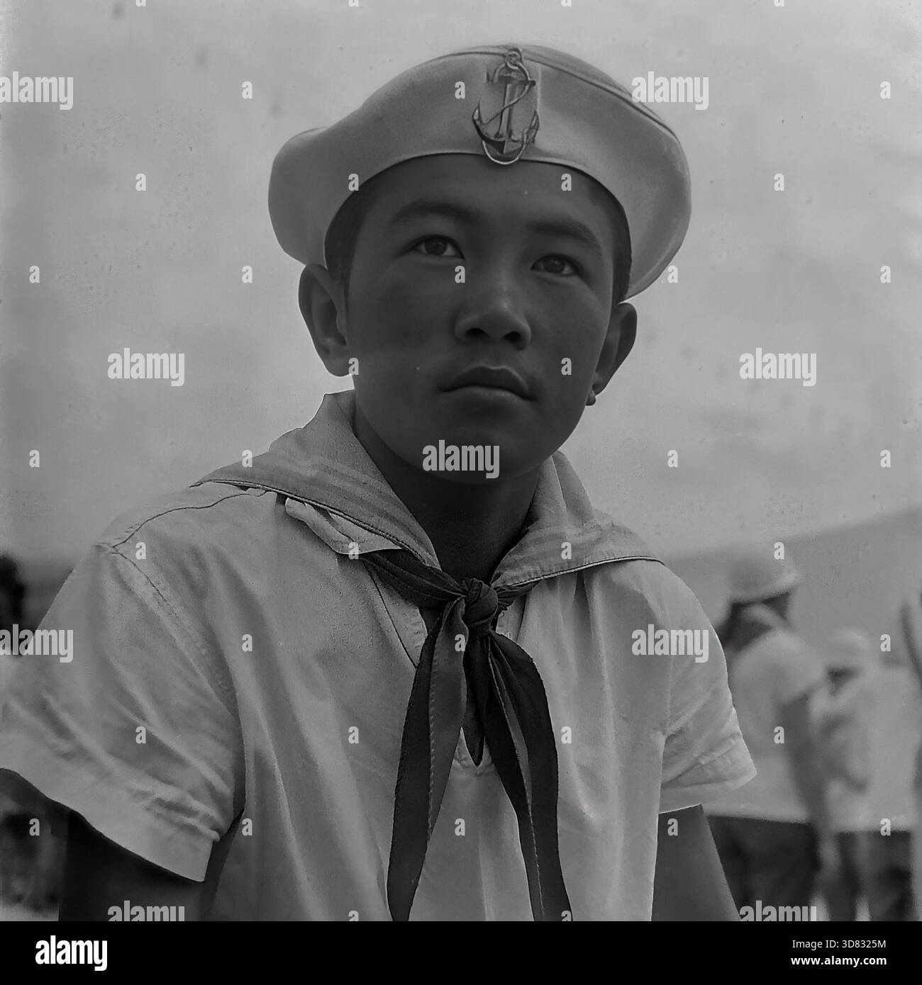 Crimée, URSS - 1968 : portrait d'un jeune garçon pionnier d'origine asiatique portant un uniforme de marin au camp international des pionniers d'Artek. Il porte une casquette blanche avec un badge ancre et un mouchoir pionnier. Le garçon regarde sérieusement au loin. En arrière-plan, d'autres enfants portant des uniformes similaires sont visibles. Cette photo d'archives en noir et blanc capture l'atmosphère multiethnique, la mode de la jeunesse soviétique et l'esprit d'amitié internationale dans la célèbre station balnéaire de Crimée pendant les vacances d'été. Banque D'Images