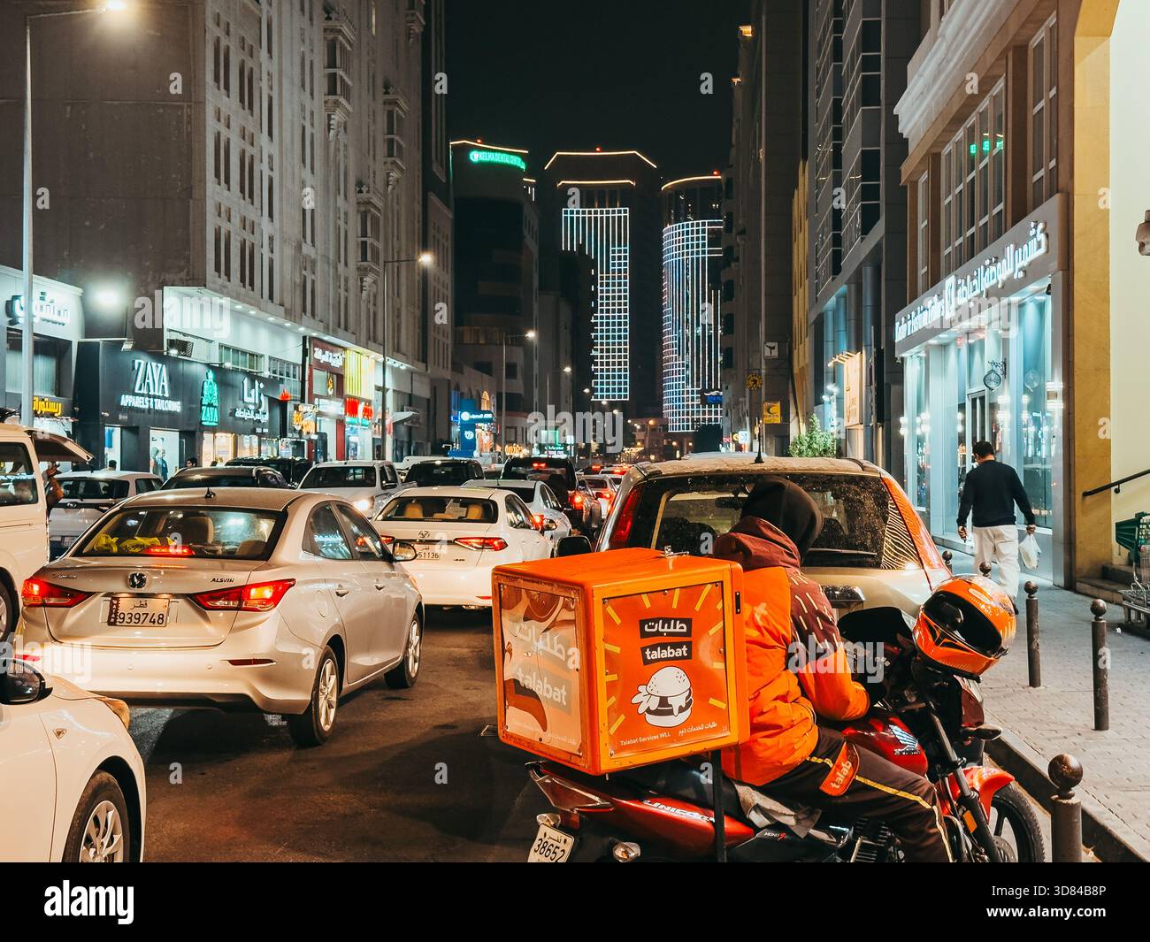 Circulation nocturne sur la rue Al Jazeera à Doha, Qatar, avec un livreur Talabat au milieu des voitures occupées et des tours de ville illuminées. Banque D'Images