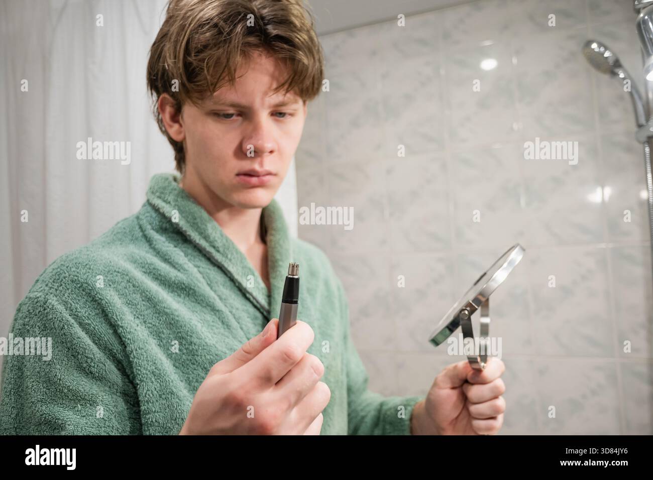 Un jeune homme caucasien portant un peignoir vert utilise une tondeuse électrique pour broyer sa pilosité faciale. Routine de rasage matinal. Gros plan. Banque D'Images