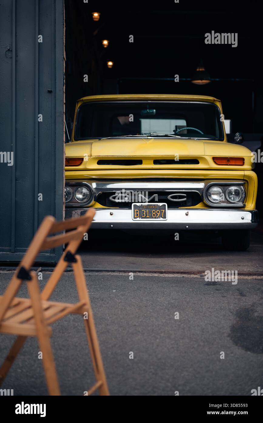 Camionnette GMC classique jaune vif des années 1960 jetant un coup d'œil à une porte de garage sombre. Une chaise pliante en bois est floue au premier plan. Banque D'Images