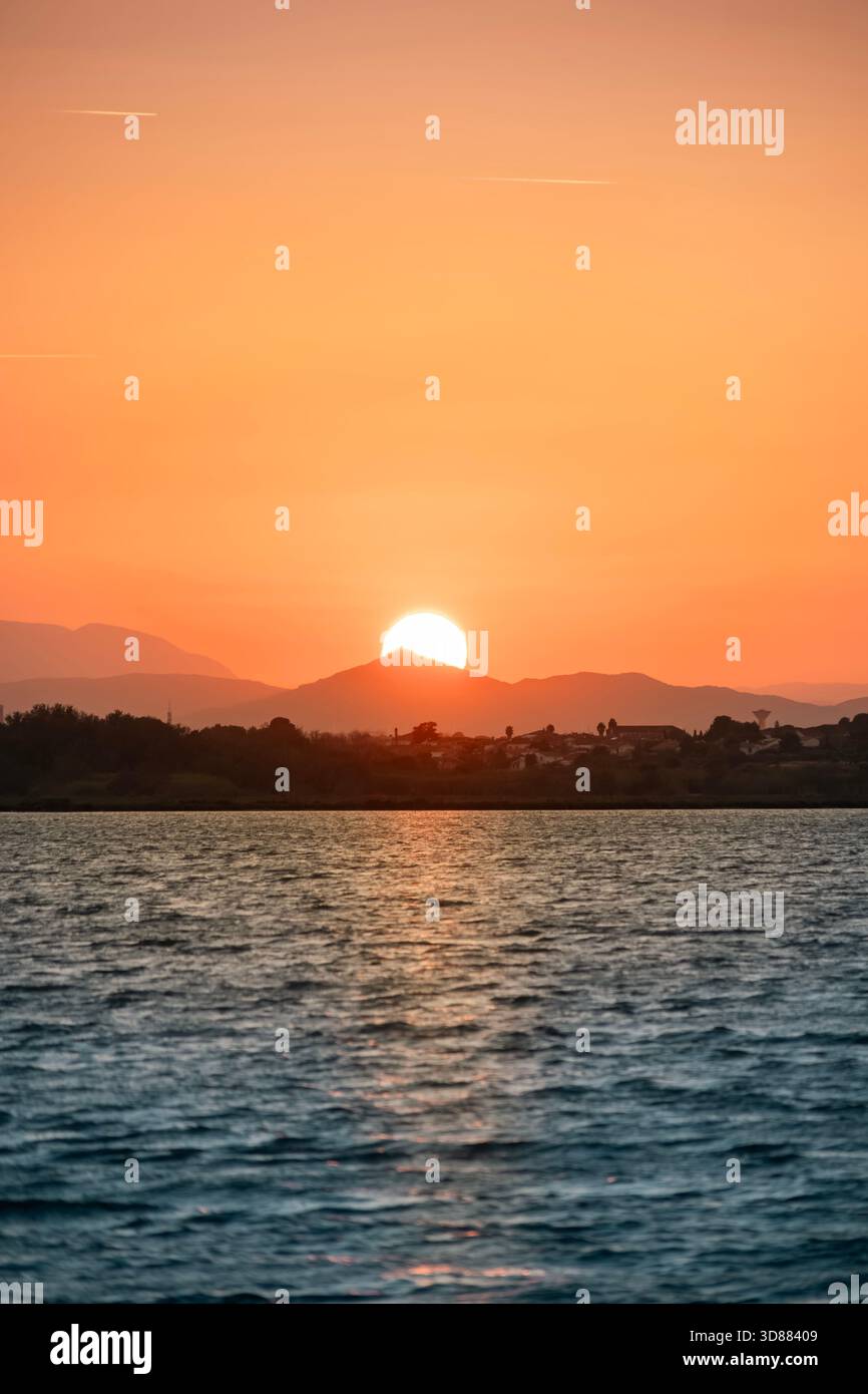 Vue verticale du coucher du soleil ou se levant sur les collines et les bâtiments côtiers, avec une lumière orange spectaculaire se reflétant sur la mer Banque D'Images