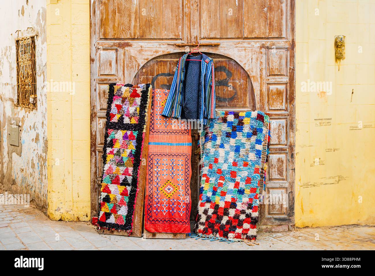 Tapis berbères colorés faits à la main et textiles traditionnels exposés contre une porte altérée à Essaouira, Maroc Banque D'Images