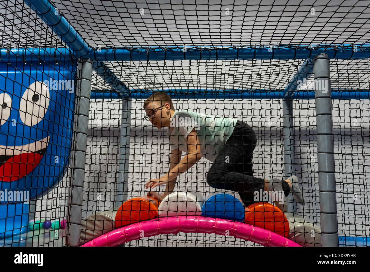 Garçon avec des lunettes rampant sur de grandes balles souples dans une structure de jeu en filet à l'aire de jeux intérieure. Loisirs actifs de l'enfance, déve de coordination physique Banque D'Images