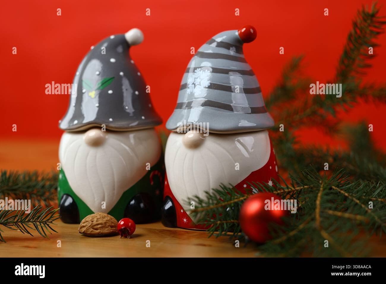 Deux décorations de gnome de Noël en céramique présentées sur un fond rouge vif, entourées de branches d'épinette et de petits ornements. Banque D'Images