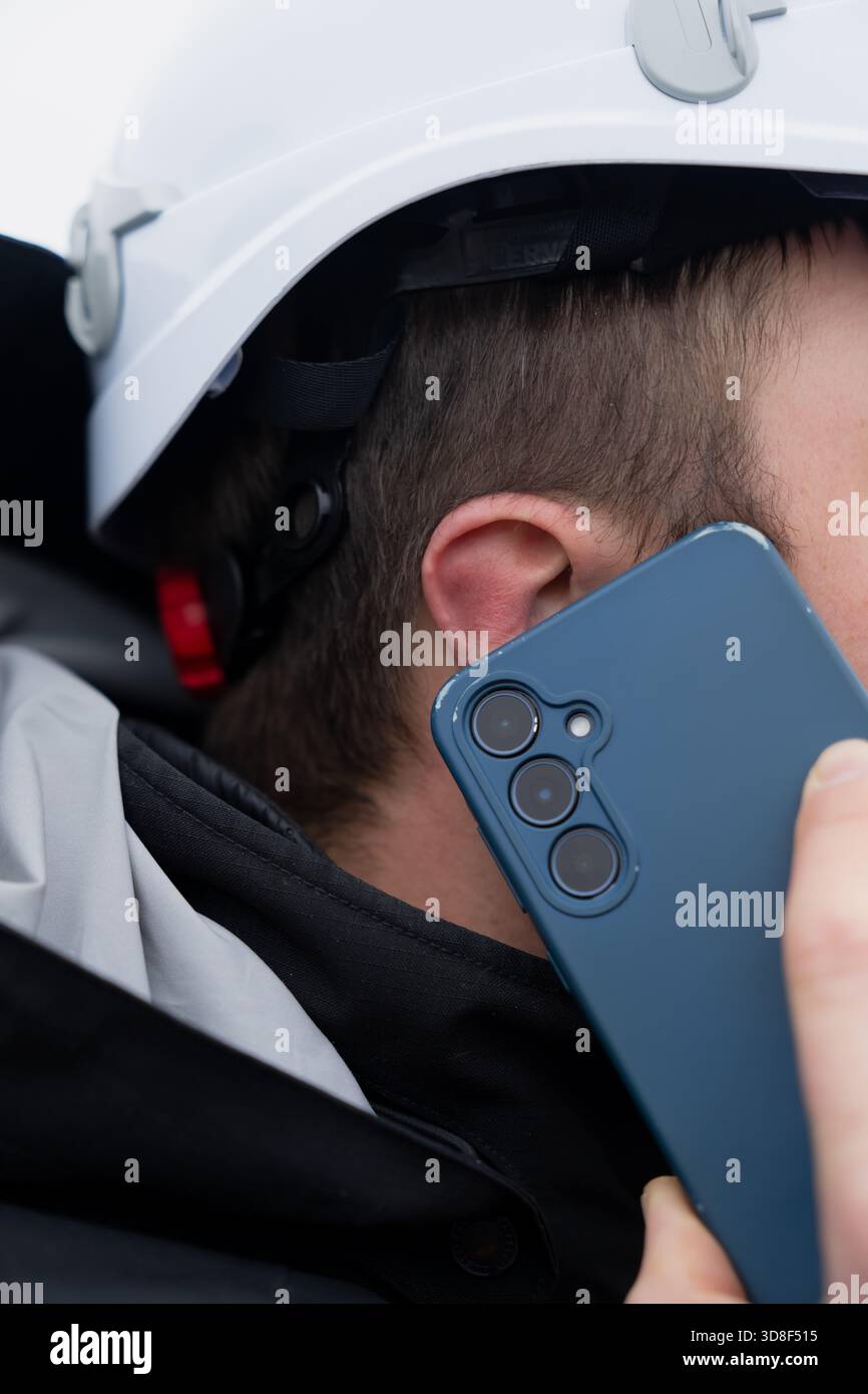 Vue détaillée d'un travailleur muni d'un casque de sécurité parlant sur un smartphone lors d'opérations sur site actives dans un environnement industriel extérieur Banque D'Images