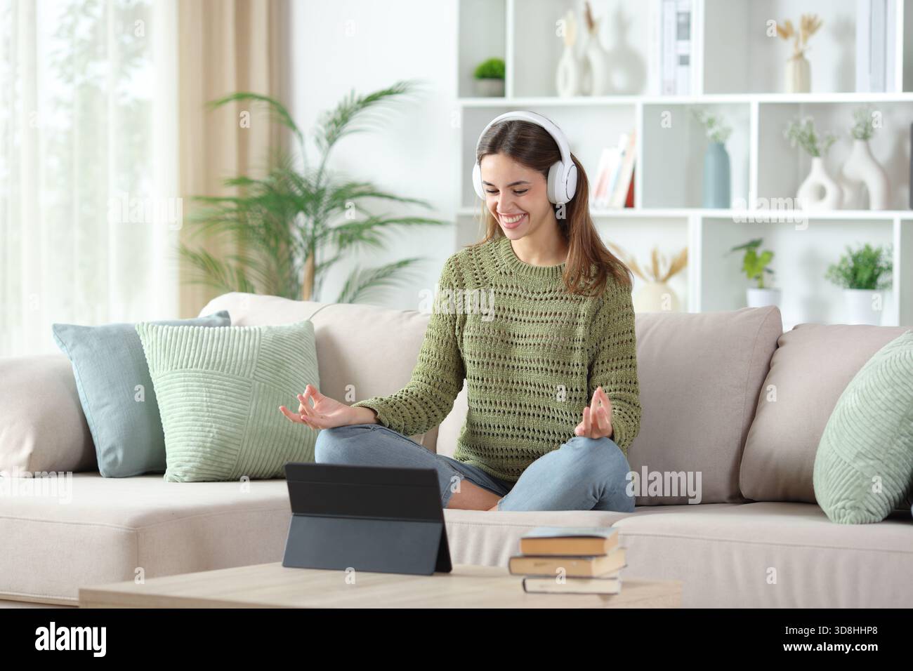 Femme heureuse en vert portant casque regardant cours de yoga en ligne sur tablette assis sur un canapé dans un intérieur de maison Banque D'Images