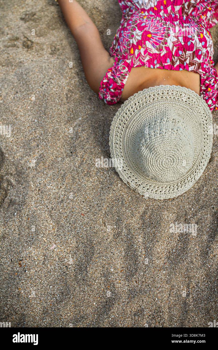 Femme asiatique en robe de plage colorée couchée en faisant une sieste sur le sable de plage couvrant son visage avec chapeau crochet rond pendant la retraite d'été et les vacances Banque D'Images